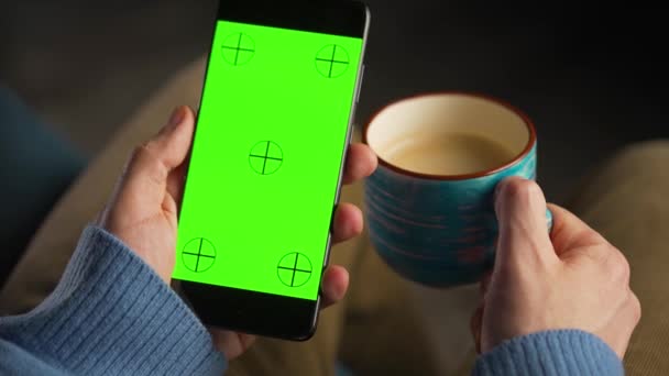 Άνθρωπος που χρησιμοποιεί smartphone με πράσινη οθόνη mock-up σε κάθετη λειτουργία και πίνοντας καφέ. Περιήγηση στο Internet, παρακολούθηση περιεχομένου, βίντεο. - Πλάνα, βίντεο
