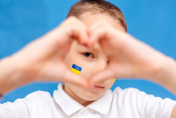 A child with a flag on his cheek. Caucausian child looks into the camera. War in Ukraine. Space for text. Patriotism. Banner. Close up. Child makes a heart with his hands on a blue background. - Фото, зображення