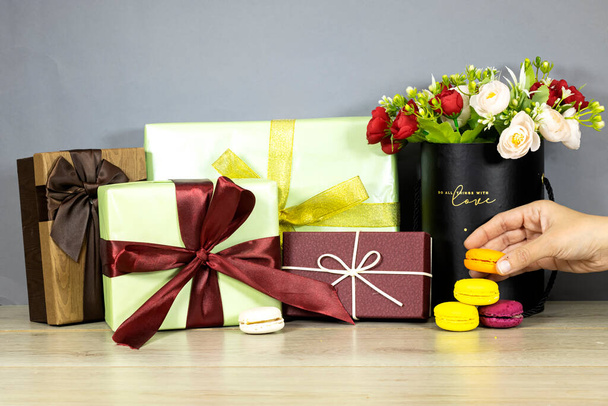 boîtes cadeaux en papier pile et bouquet de fleurs dans une boîte noire et tenir à la main des biscuits de macaron français colorés sur une table en bois. Festive Noël et Nouvel An fête concept de célébration - Photo, image