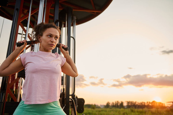 Determined active athletic middle aged woman exercising on an outdoor exercise machine, doing arm exercises at sunset - Zdjęcie, obraz