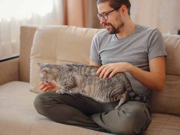 owner plays with cat while relaxing on sofa in living room. A young man is resting with pet at home. Pets concept - Photo, Image