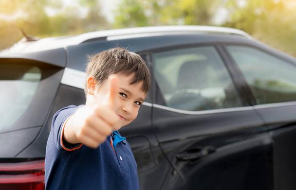 Onnellinen nuori poika osoittaa peukalo ylös, kun stnading auton vieressä, koulun lapsi hymyilevät kasvot seisoo parkkipaikalla aurinkoisena päivänä Kevät tai kesä, Positiivinen lapsi menee kouluun. - Valokuva, kuva