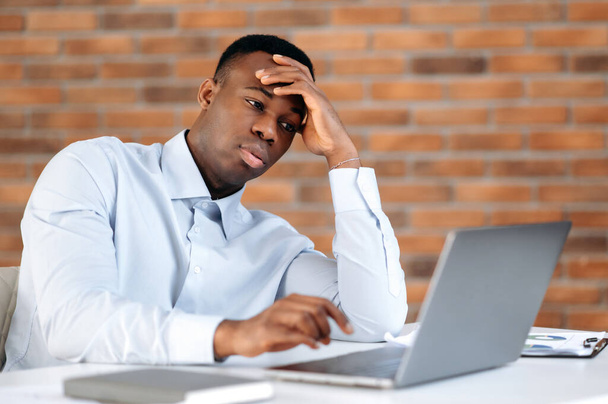 Upset bored african american man, freelancer or IT specialist, works in the office, sitting at a desk with laptop, overworked from online work, tired, have a stressful situation, need a rest - 写真・画像