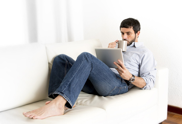 Jovem atraente homem hispânico em casa no sofá branco usando tablet digital ou pad
 - Foto, Imagem