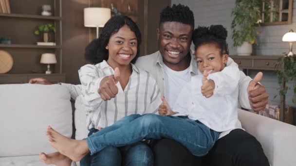 Happy African family young guardian parents and little daughter adopted girl child sitting on sofa at home own apartment new house looking at camera show thumbs up approve recommend positive attitude - Filmati, video