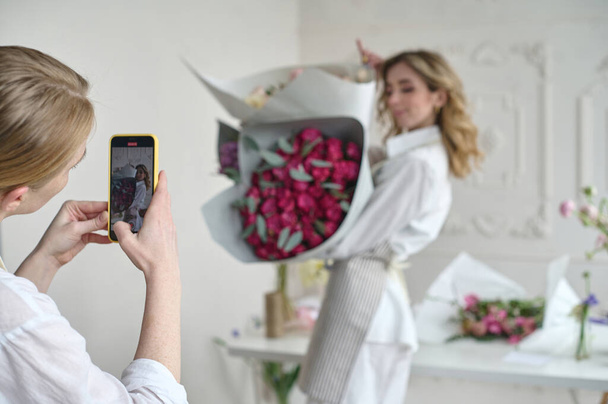 two beautiful young florists in aprons with bouquets in their hands taking a selfie on a white background - Фото, зображення