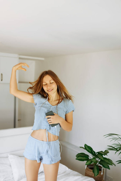 Young teen girl in pajama standing on her bed and singing a song. Cheerful caucasian adolescent girl with headphones dancing on her bedroom  - Valokuva, kuva