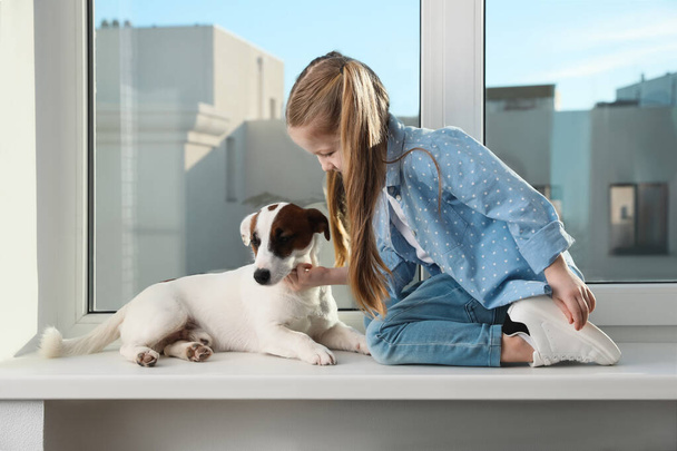 Cute little girl with her dog on window sill indoors. Childhood pet - Foto, Bild