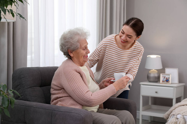 Young caregiver giving tea to senior woman in room. Home care service - Φωτογραφία, εικόνα