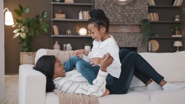 Happy relaxing african mother lying on sofa and small child active playful daughter girl on couch laughing having fun playing talking conversation smiling mum enjoying bonding spending time with kid - Кадры, видео