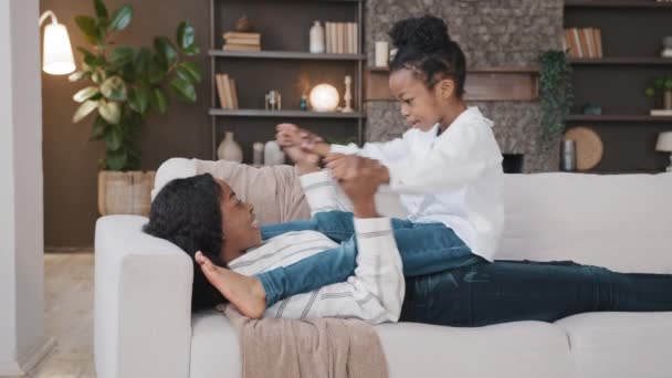 African American loving mother mommy lying on sofa relaxing at home living room with little daughter child talking having fun casual game playing holding hands baby rocking back forward sitting on mum - Footage, Video