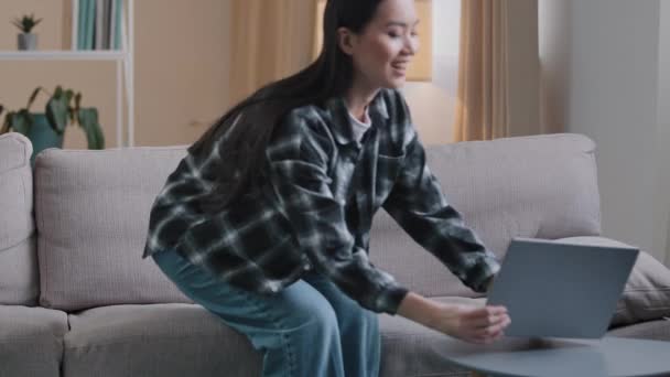 Carefree Asian girl enters living room sit down on comfortable sofa takes laptop with smile and watches video online woman uses computer sitting on couch at home casual browsing net resting typing - Metraje, vídeo