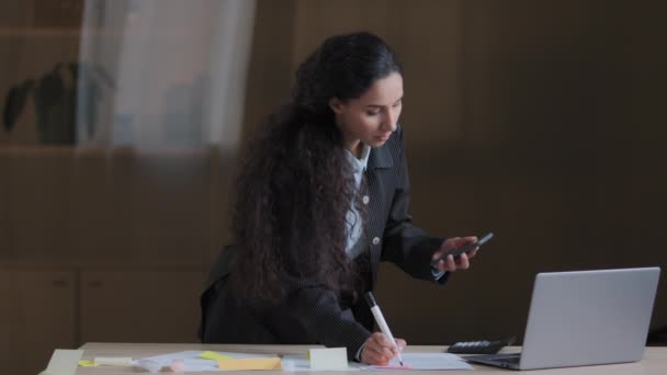 Сосредоточенная женщина арабский работник дизайнер бизнес-женщина, стоящая за столом офиса с помощью мобильного телефона проверки информации написания задач на липкие заметки организации работы управления документами план проекта - Кадры, видео