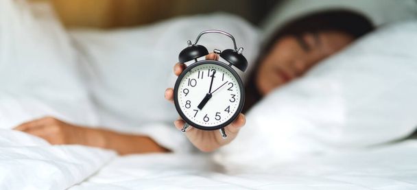 Una mujer asiática sosteniendo y mostrando un reloj despertador mientras duerme en una cama blanca y acogedora por la mañana - Foto, Imagen