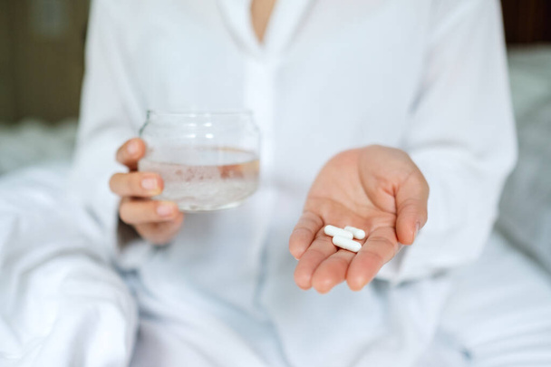 Closeup image of a sick woman holding white pills and a glass of water while sitting on a bed  - Photo, Image