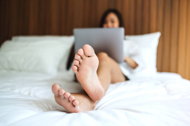 Une belle femme asiatique utilisant et travaillant sur un ordinateur portable tout en étant couché sur un lit confortable blanc à la maison - Photo, image