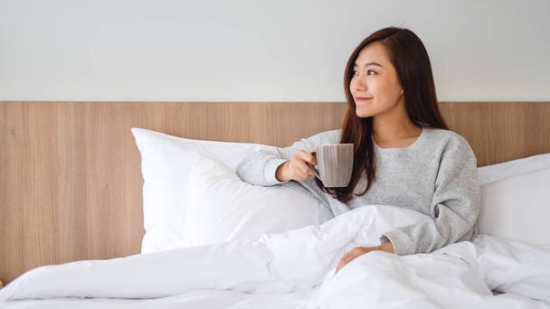 午前中に家で白い居心地の良いベッドの上に横たわっている間に熱いコーヒーを飲む美しいアジアの女性 - 写真・画像
