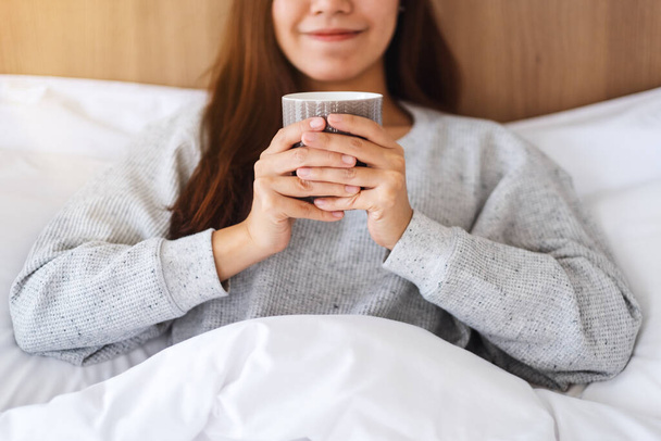 Μια όμορφη Ασιάτισσα πίνει ζεστό καφέ ενώ ξαπλώνει σε ένα λευκό άνετο κρεβάτι στο σπίτι το πρωί - Φωτογραφία, εικόνα