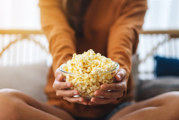 Close up image of a woman holding a bowl of pop corn while sitting on sofa at home - Фото, изображение
