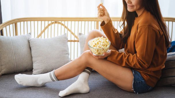Close up image of a beautiful young woman holding and eating pop corn while sitting on sofa at home - Photo, image