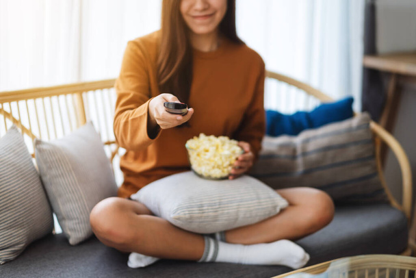 De cerca la imagen de una hermosa mujer joven comiendo maíz pop y canal de búsqueda con control remoto para ver la televisión mientras está sentado en el sofá en casa - Foto, Imagen