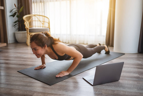 Eine schöne junge Asiatin macht Liegestütze auf der Trainingsmatte, während sie zu Hause Online-Workout-Tutorials auf dem Laptop anschaut - Foto, Bild