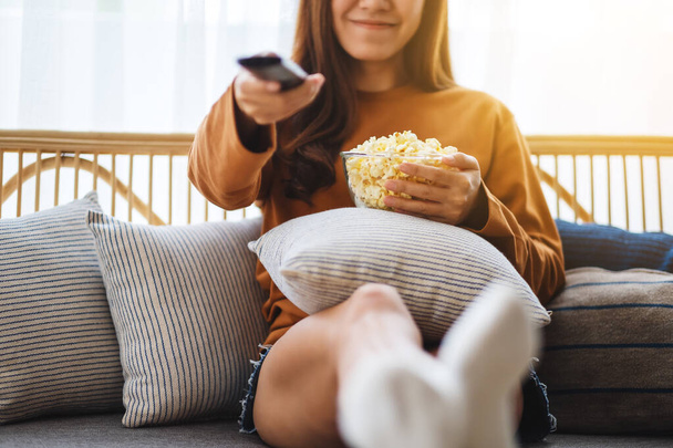 Nahaufnahme Bild einer schönen jungen Frau, die Popcorn isst und Kanal mit Fernbedienung sucht, um fernzusehen, während sie zu Hause auf dem Sofa sitzt - Foto, Bild