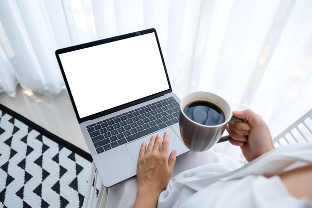 Макет жінки, яка працює і друкує на ноутбуці з порожнім екраном, сидячи і п'ючи каву в спальні вдома
 - Фото, зображення
