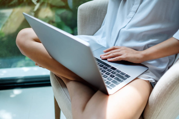 Detailní záběr ženy pracující na notebooku, zatímco sedí na bílém křesle doma se zeleným přírodním pozadím - Fotografie, Obrázek