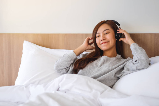 Μια όμορφη Ασιάτισσα απολαμβάνει να ακούει μουσική με ακουστικά ενώ βρίσκεται σε ένα λευκό άνετο κρεβάτι στο σπίτι  - Φωτογραφία, εικόνα