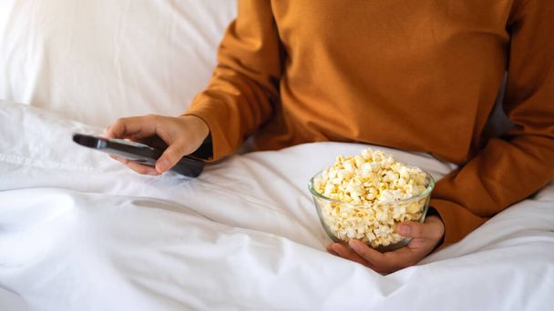 Close up image of a woman eating pop corn and searching channel with remote control to watch tv while sitting in bed - Фото, изображение