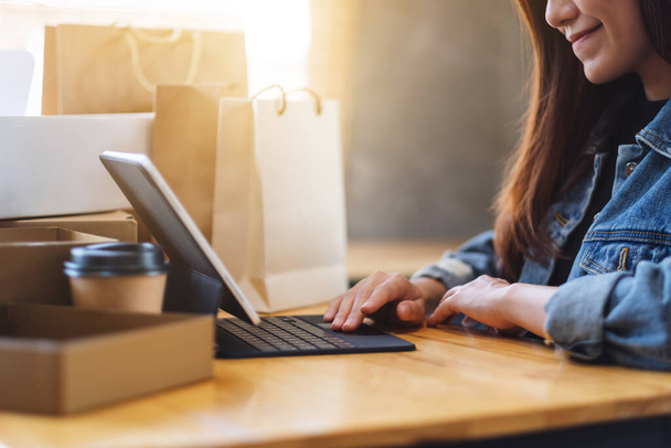 Szoros kép egy fiatal ázsiai nő használja tabletta pc online vásárlás postaláda és bevásárló táskák az asztalon otthon - Fotó, kép