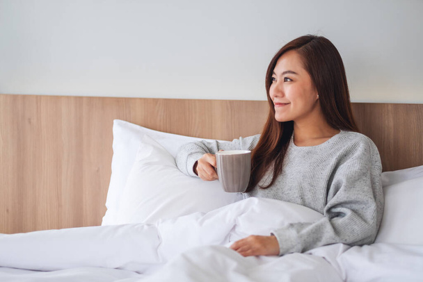 午前中に家で白い居心地の良いベッドの上に横たわっている間に熱いコーヒーを飲む美しいアジアの女性 - 写真・画像