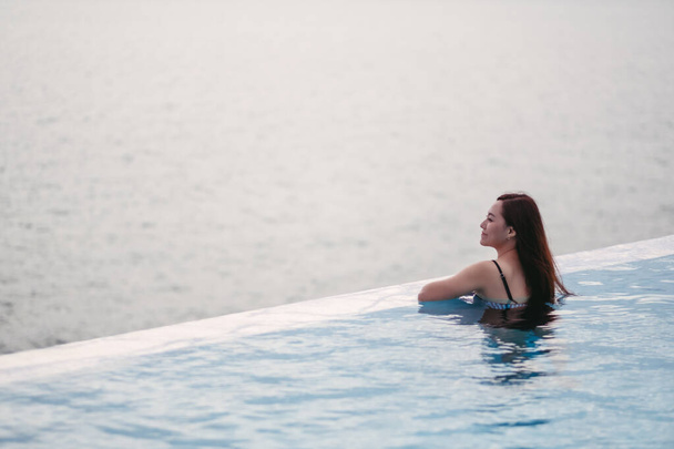 Eine junge asiatische Frau entspannt sich im Infinity-Pool und blickt auf einen wunderschönen Meerblick - Foto, Bild