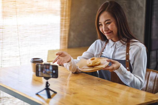 A beautiful asian woman food blogger or vlogger showing a piece of donut while recording a video on camera  - Foto, imagen