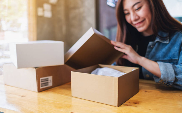 A beautiful young asian woman receiving and opening a postal parcel box at home for delivery and online shopping concept - 写真・画像