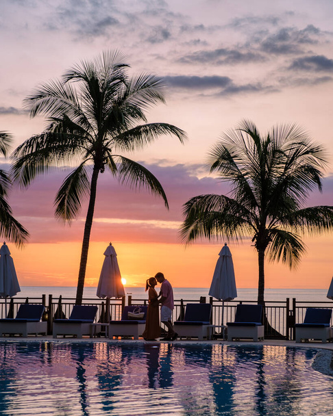couple man and woman watching sunset by a swimming pool with tropical palm trees during a luxury vacation holiday - Zdjęcie, obraz