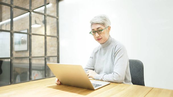 maduro ásia negócios mulher usa óculos usando laptop computador sentar no local de trabalho mesa. feliz sênior mais velho empregado 50s empresário - Foto, Imagem