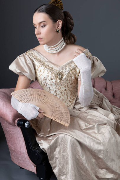 A Victorian woman wearing a gold ball gown and sitting on a pink antique sofa - Photo, Image