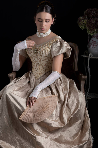 A Victorian woman wearing a gold ball gown and sitting on an antique chair against a dark backdrop - Foto, Imagem