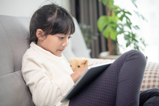 Little girl using tablet playing game on the internet, Kid sitting on sofa watching or talking with a friend online, Child relaxing in living room in the morning, Children with New Technology concept - 写真・画像