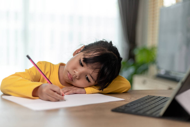 Little Asian girl sitting alone and looking out with a bored face, Preschool child laying head down on the table with sad  bored with homework, spoiled child - Photo, Image