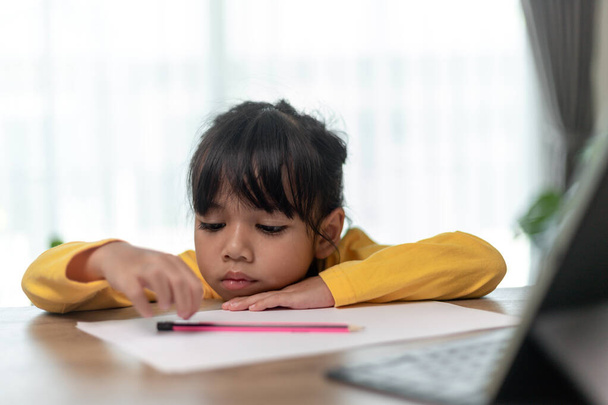 Kleines asiatisches Mädchen, das allein sitzt und mit gelangweiltem Gesicht nach draußen schaut, Kindergartenkind, das den Kopf auf den Tisch legt, traurig gelangweilt von den Hausaufgaben, verwöhntes Kind - Foto, Bild