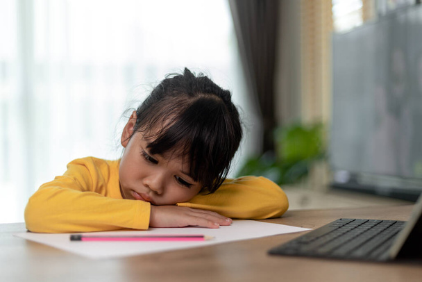 Kleines asiatisches Mädchen, das allein sitzt und mit gelangweiltem Gesicht nach draußen schaut, Kindergartenkind, das den Kopf auf den Tisch legt, traurig gelangweilt von den Hausaufgaben, verwöhntes Kind - Foto, Bild