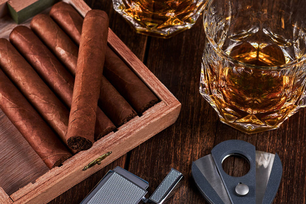 Caja con puros cubanos, encendedor y cortador en la mesa de madera vieja. Dos vasos de whisky o alcohol en el fondo. - Foto, imagen
