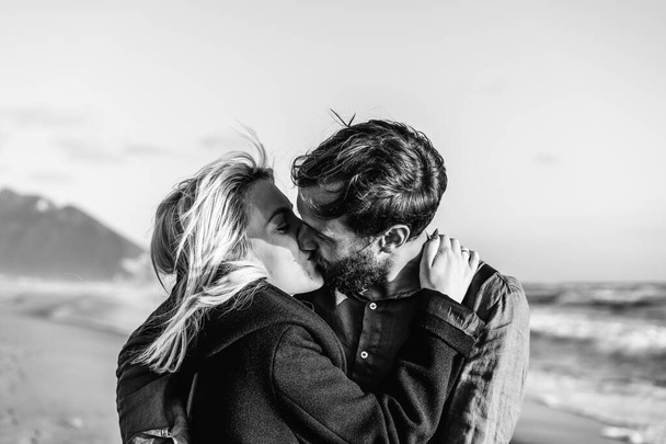 Romantic hipster couple in love kissing on shoreline at the wild beach - Boyfriend and girlfriend wearing cool clothing and vintage backpack kissing in front of the sea during the sunset - Black and white - Photo, Image