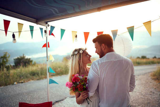 Newlywed couple looking at each other in front of decorated camper. Boho wedding. Wedding ceremony, love, nature concept - Foto, Bild