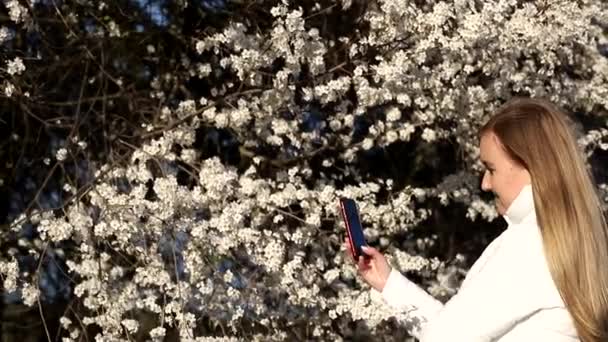Ragazza bionda in una maschera bianca fa un selfie su uno sfondo di alberi da frutto in fiore - Filmati, video