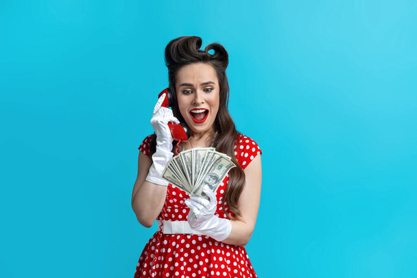 Excited young pinup woman in red polka dot dress holding money, speaking on landline phone on blue studio background - Fotografie, Obrázek
