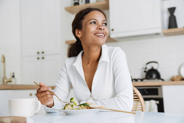 Mladá černošky žena s úsměvem při obědě v kuchyni doma - Fotografie, Obrázek